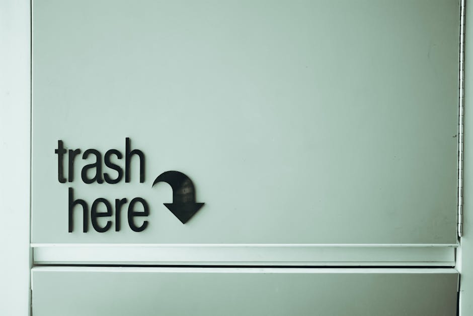 A close-up view of a light grey wall with a black, three-dimensional sign that reads 'trash here' in lowercase letters, accompanied by a downward-pointing arrow. The sign is mounted on a smooth, matte surface, and the arrow suggests the location of a rubbish bin or collection point. The wall appears clean and indoors, possibly within a waste management or disposal area, with minimal visible environmental details. The overall scene reflects a designated space for rubbish collection, relevant to private waste handling or alternative rubbish removal services such as those offered by Rubbish Clearance Streatham, contrasting with standard local authority collection points. The lighting is even, highlighting the clarity and simplicity of the signage and wall surface.
