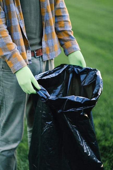A person wearing a yellow and gray checkered shirt, with a green long-sleeved top underneath, and green gloves, is holding open a large black plastic rubbish bag outdoors on a grassy area. The individual appears to be preparing for waste collection or clearance, with the bag positioned for loading waste materials. The background features a blurred view of green grass, indicating a park or garden environment. The person's lower body is visible, showing beige trousers and a brown belt. This scene exemplifies a private waste handling activity, possibly associated with independent rubbish removal services, emphasizing the use of durable black plastic bags for waste collection in an outdoor setting, highlighting the importance of proper rubbish containment during clearance tasks conducted by companies such as Rubbish Clearance Streatham.
