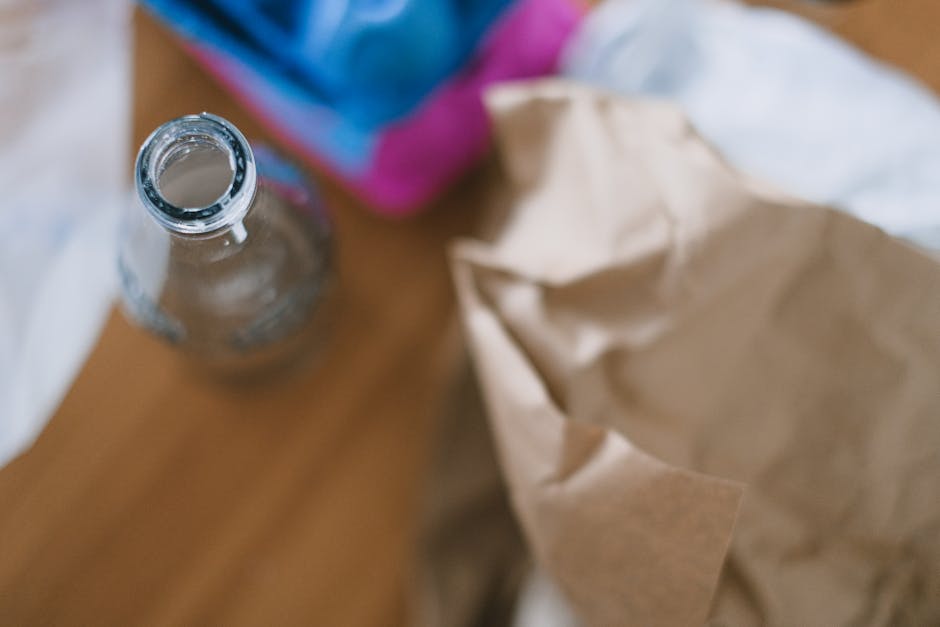 A clear glass bottle with a narrow neck and open top is positioned on a wooden surface in the foreground, showing a smooth, transparent texture typical of glass. Behind the bottle, there is a partially visible cardboard box or paper bag with a crumpled, light brown surface, indicating it is made from kraft paper or cardboard. To the upper left, a portion of a fabric item with a vibrant pink and blue color pattern is visible, adding a splash of colour to the scene. The background appears slightly out of focus but hints at a cluttered environment, possibly a home or storage area, with various items and surfaces. The lighting is natural and soft, illuminating the objects without harsh shadows. This scene exemplifies typical clutter associated with household waste or rubbish awaiting collection, highlighting the types of rubbish that might be handled during a private waste clearance service, such as those offered by Rubbish Clearance Streatham.