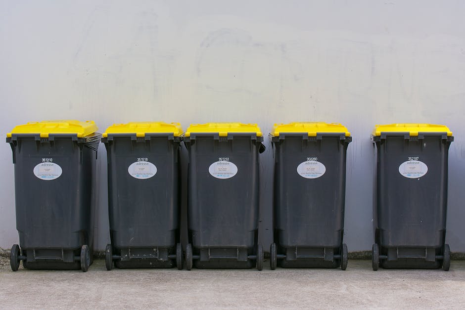 A row of five large, black wheeled rubbish bins with yellow lids positioned against a plain, light-colored wall in an outdoor setting. Each bin features a white oval label with barcode and serial number details, indicating they are part of a waste collection system for private disposal or alternative waste handling. The bins are aligned on a concrete surface, with the first three showing slight wear and dirt, while the remaining two appear cleaner. The lighting is natural, casting soft shadows on the ground and wall, emphasizing the textured surfaces of the bins and the smooth finish of the wall behind them. The scene reflects a typical on-site waste management arrangement used for rubbish clearance, likely arranged through a private service provider such as Rubbish Clearance Streatham, suitable for on-demand rubbish removal or managed waste disposal outside standard municipal collection services.