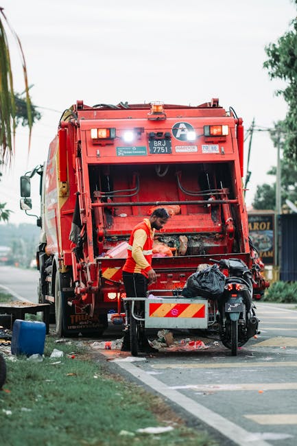 A street scene showing a waste collection worker standing behind a large red rubbish truck with the rear loading mechanism extended and open, indicating active waste pickup. The worker, wearing a yellow high-visibility vest over a red shirt, is handling a black plastic bag placed on a small metal platform attached to the truck. The truck is positioned on the roadside, with its rear facing the sidewalk, and appears to be actively engaged in waste collection, possibly as part of private rubbish removal services. Surrounding the scene, there is a patch of grass and a paved road extending into the distance, lined with utility poles and trees, with some buildings and signs visible further down. The environment is outdoor during daylight, with a slightly overcast sky, contributing to even lighting without harsh shadows. The scene reflects typical roadside waste management activity aligned with independent rubbish clearance and disposal tasks performed outside of local authority services, as managed by companies like Rubbish Clearance Streatham.