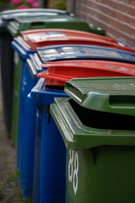 A row of large wheelie bins lined up against a brick wall on a paved pathway, with the nearest green bin in the foreground and additional bins in blue, red, and dark green extending into the background. The green wheels and textured lids, with some partially open, are visible, along with white markings on the side of the bins. The scene suggests an outdoor setting for waste storage, typical of private rubbish collection points, with the bins organized neatly and aligned along the edge of the pavement, naturally associated with domestic or commercial rubbish removal services provided by companies like Rubbish Clearance Streatham. The environment appears clean and orderly, with soft natural lighting illuminating the scene without harsh shadows, emphasizing the different colors and textures of the bins, which are made of durable plastic materials suitable for waste disposal. This image illustrates a typical rubbish storage arrangement often used for independent waste collection efforts as part of on-site clearance or private disposal services.